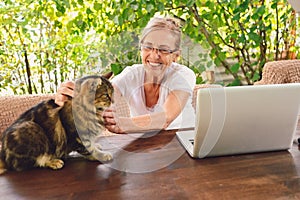 Happy senior woman with home cat use wireless headphones working online with laptop computer outdoor