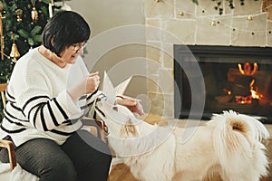 Happy senior woman playing with adorable white dog in stylish festive christmas living room. Beautiful woman enjoying cozy