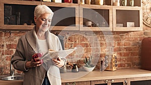 Happy Senior Woman Reading Newspaper And Drinking Hot Tea In Kitchen