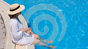 happy senior woman relaxing near blue outdoor swimming pool with blue cocktail wearing straw hat. People are enjoying