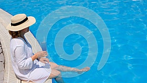 happy senior woman relaxing near blue outdoor swimming pool with blue cocktail wearing straw hat. People are enjoying