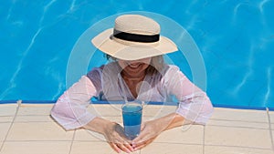 happy senior woman relaxing near blue outdoor swimming pool with blue cocktail wearing straw hat. People are enjoying