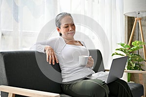 Happy senior woman sipping coffee while working on her business tasks on laptop