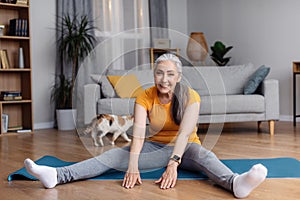 Happy senior woman stretching sitting on yoga mat, doing flexibility exercises, making domestic workout in living room
