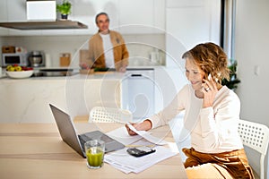 Happy senior woman talking on smartphone and checking bills when husband preparing dinner at kitchen, selective focus