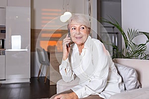 Happy senior woman using mobile phone at home. Smiling cool old woman with white hair sitting on sofa and messaging with