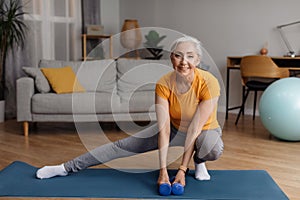 Happy senior woman warming up, doing legs stretching exercises while working out at home on fitness mat