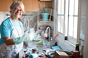 A happy senior woman while wash dishes. Gray hair and eyeglasses. wearing apron and gloves. Corner of kitchen. Grandmother at work