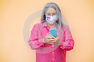 Happy senior woman wearing protective face mask using mobile phone outdoor - Focus on face