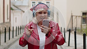 Happy senior woman in wireless headphones choosing, listening music in smartphone dancing outdoors