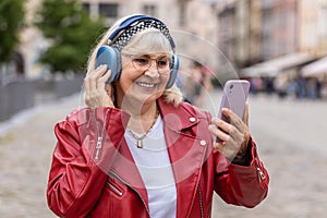 Happy senior woman in wireless headphones choosing, listening music in smartphone dancing outdoors