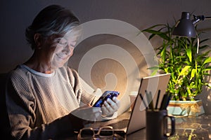 Happy senior woman working and using laptop at late night, looking at mobile phone message