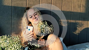 Happy and sensual woman taking selfie photo on mobile phone with flowers while sitting on bed at home