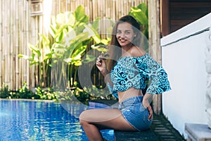 Happy sexy girl in denim shorts and top sits on pool. Young tanned model resting on summer day