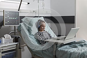 Happy sick little girl under treatment sitting on patient bed with laptop