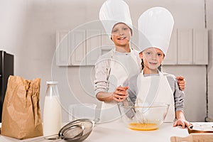 happy sister and brother in aprons and chef hats whisking eggs in bowl at table