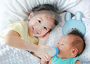 Happy sister feeding milk from bottle for her brother and lying on the bed