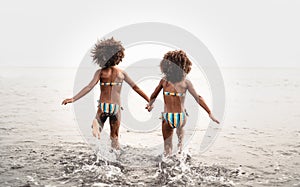 Happy sisters running inside water during summer time - Afro kids having fun playing on the beach