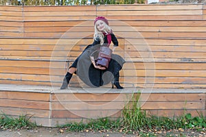 Happy sits on a bench of a summer theater, wooden in a burgundy coat and biret, looking at the camera, in the fall