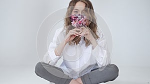 Happy sitting young girl puts on mask of fresh flowers, breaths clean air indoor