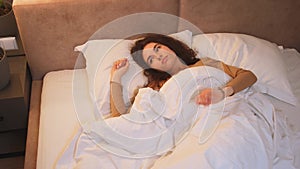 Happy sleepy young adult woman waking up lying in comfortable white bed. Positive pretty girl stretching enjoying early