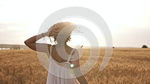 Happy, slender girl in straw hat and summer white dress happily running in clear wheat field. Sun shines on the