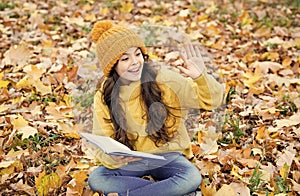 Happy small child with school book give salute with hand sitting on autumn leaves outdoors, literature