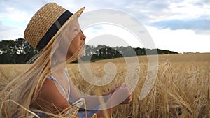 Happy small child in straw hat holding cereal spikelet in hands while sitting in the wheat field. Beautiful little girl