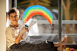 Happy small european boy chooses paints, draws rainbow on window in children room interior