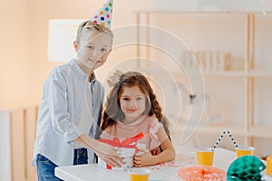 Happy small girl and boy hold gift box with red ribbon, prepare surprise on birthday, pose at white table, wear cone party hats,