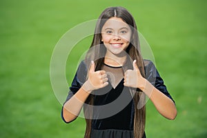 Happy small girl child with long hair and beauty look wear fashion dress showing thumbs ups hands gesture green grass
