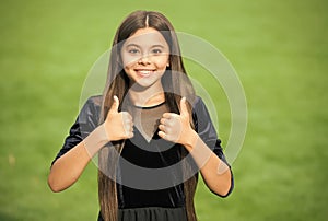 Happy small girl child with long hair and beauty look wear fashion dress showing thumbs ups hands gesture green grass