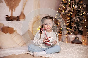 Happy small girl holding traditional Christmas striped candy cane on lights background at home. Smiling Little girl sitting on bed