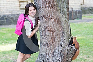 Happy small kid in uniform with school backpack resarch squirrel climbing tree in park, zoology.