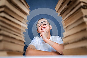 Happy smart boy in glasses sitting between two piles of books an