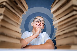 Happy smart boy in glasses sitting between two piles of books and look up smiling