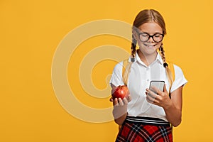 Happy smart caucasian teen girl student in glasses hold red apple, typing on phone