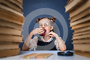 Happy smart girl in rounded glasses, eating a red apple while sitting between two piles of books and look at camera