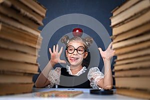 Happy smart girl in rounded glasses with red apple on her head sitting between two piles of books, have fun and look at