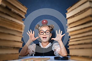 Happy smart girl in rounded glasses with red apple on her head sitting between two piles of books, have fun and look at