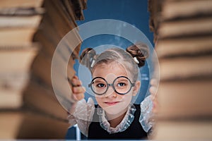 Happy smart girl in rounded glasses sitting between two piles of books and look at camera smiling