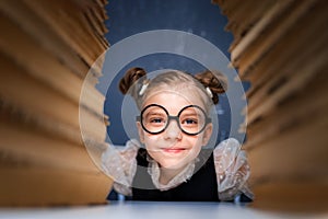 Happy smart girl in rounded glasses sitting between two piles of books and look at camera smiling