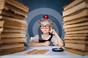 Happy smart girl in rounded glasses thoughtfully sitting between two piles of books with red apple on head, pointing