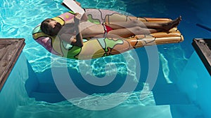 Happy smart girl sunbathing on inflatable mattress in swimming pool reading book in sunbeam. Wide shot of relaxed