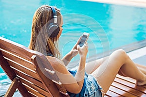 Happy smartphone woman relaxing near swimming pool listening with earbuds to streaming music. Beautiful girl using her mobile phon