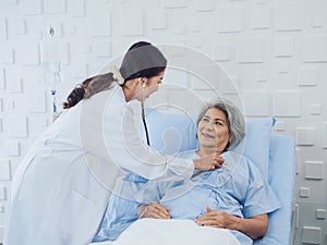 Happy smile Asian elderly old female patient in light blue dress lying on bed while beautiful young woman doctor.