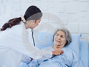 Happy smile Asian elderly old female patient in light blue dress lying on bed while beautiful young woman doctor.