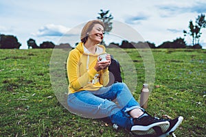Happy smile girl holding in hands cup of hot tea on green grass in outdoors nature park, beautiful young woman hipster enjoy drink