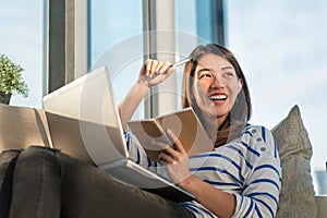 Happy smile young asian woman working with laptop and hold pen with notebook and sitting on sofa at home office, Selective focus