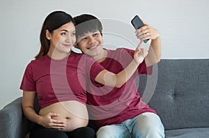 Happy smiley young couple dressed casually sitting on a couch together smiling and doing a selfie. They are expecting a healthy